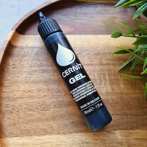 May include: A black tube of Cernit Gel, a liquid hardener for polymer clay, with a white drop logo and text. The tube contains 1 fl oz (30 ml) of gel and is made in Belgium. The tube is on a wooden tray.