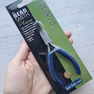 Può includere: Un paio di pinze a becco curvo a catena con manico blu e codice colore. La confezione presenta il testo "The Bead Smith Pliers" e "Color 1.0."