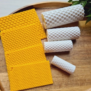 Può includere: Un vassoio di legno contiene diversi fogli gialli a motivo a nido d'ape e candele di cera d'api bianche arrotolate. Le candele variano di dimensioni e i fogli sono di un giallo vibrante. Le foglie verdi aggiungono un tocco naturale.