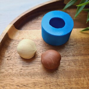 Pode incluir: Duas pequenas bolas de argila, uma bege e outra marrom, sobre uma superfície de madeira ao lado de uma forma de silicone azul.