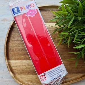 Puede incluir: Un paquete de arcilla de modelar FIMO Soft roja, horneable, con un peso de 454 g (1lb). El paquete es blanco con detalles azules y rosas, y la marca "FIMO" es visible. La arcilla está sobre una bandeja de madera.