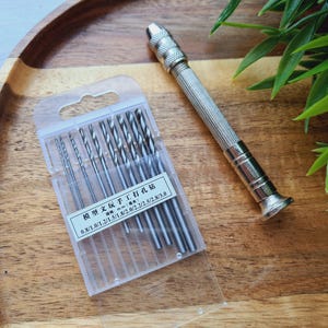 Puede incluir: Un juego de brocas plateadas en una funda de plástico transparente, junto a un taladro manual plateado. Las brocas varían en tamaño de 0,8 mm a 3,0 mm. El texto en la funda dice "Model Wenwan Manual Punching Drill".