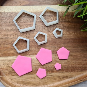May include: A set of six white plastic hexagon cookie cutters and six pink fondant hexagons of varying sizes on a wooden surface.
