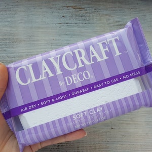Peut inclure: Un paquet de pâte à modeler souple Claycraft by Deco, à rayures violettes et blanches. L'emballage indique "Air Dry, Soft & Light, Durable, Easy to Use, No Mess". L'emballage indique également "Soft Clay" et "Net Wt 137 g (4.8 oz)".