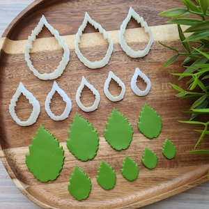 May include: A set of 10 white plastic leaf-shaped cookie cutters and 8 green fondant leaves on a wooden tray. The cutters are in various sizes and shapes.