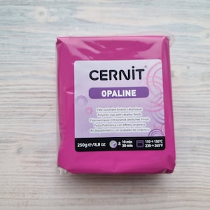 Może przedstawiać: Różowe opakowanie gliny polimerowej CERNIT OPALINE z ceramicznym wykończeniem. Opakowanie jest oznaczone instrukcjami dotyczącymi wypiekania gliny w temperaturze 110-130 stopni Celsjusza lub 230-265 stopni Fahrenheita. Opakowanie zawiera 250 gramów gliny.