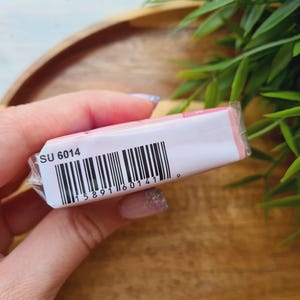 May include: A small, rectangular product wrapped in clear plastic. The packaging is white with pink accents and a barcode. The text "SU 6014" is printed on the packaging. The product is held in a person's hand.