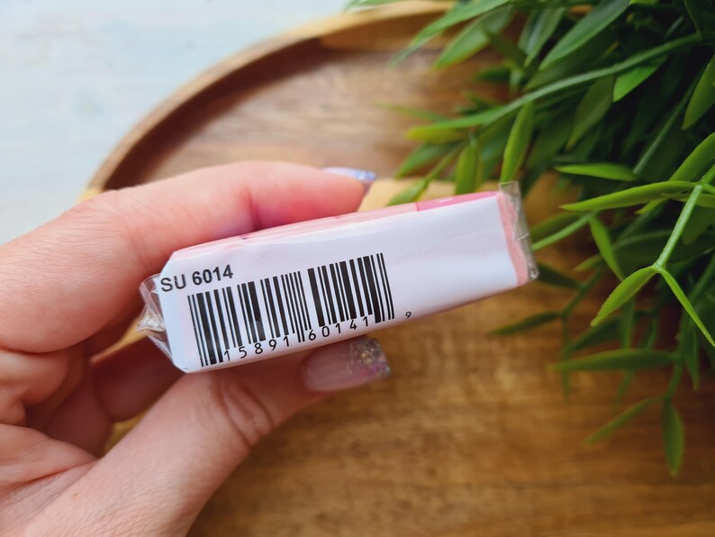 May include: A small, rectangular product wrapped in clear plastic. The packaging is white with pink accents and a barcode. The text "SU 6014" is printed on the packaging. The product is held in a person's hand.