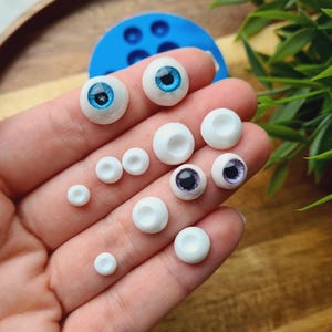 K&ouml;nnte beinhalten: Eine Sammlung von wei&szlig;en und blauen Puppenaugen in verschiedenen Gr&ouml;&szlig;en. Die Augen sind auf einer Hand angeordnet, wobei die gr&ouml;&szlig;eren Augen oben und die kleineren Augen unten liegen.