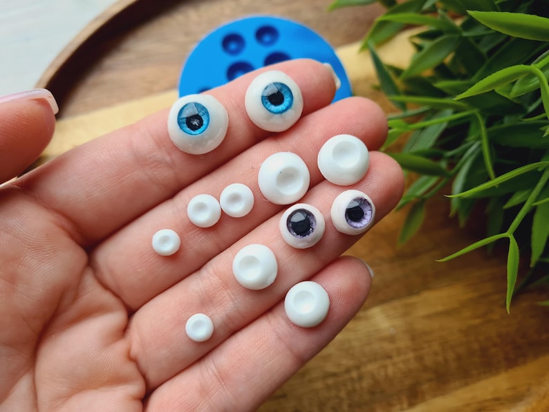 K&ouml;nnte beinhalten: Eine Sammlung von wei&szlig;en und blauen Puppenaugen in verschiedenen Gr&ouml;&szlig;en. Die Augen sind auf einer Hand angeordnet, wobei die gr&ouml;&szlig;eren Augen oben und die kleineren Augen unten liegen.