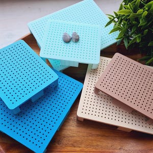 May include: An assortment of colorful pegboards in shades of blue, teal, and tan. The pegboards are square and have small holes throughout. Two small, gray stones rest on one of the teal boards. The pegboards are arranged on a wooden tray.