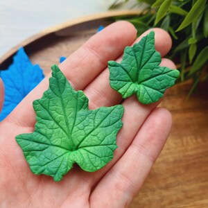 Puede incluir: Dos hojas verdes de arcilla polim&eacute;rica con un patr&oacute;n de venas detallado. Las hojas se sostienen en una mano contra un fondo de madera.