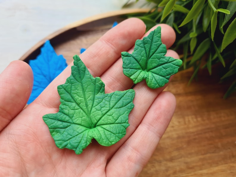 Puede incluir: Dos hojas verdes de arcilla polim&eacute;rica con un patr&oacute;n de venas detallado. Las hojas se sostienen en una mano contra un fondo de madera.