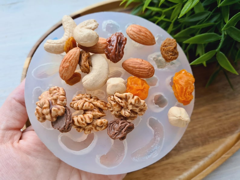 May include: A clear silicone mold with 12 cavities shaped like various nuts and dried fruits. The mold is filled with miniature replicas of almonds, cashews, walnuts, and raisins.