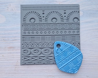 Pode incluir: Um carimbo de silicone cinza com uma variedade de padrões geométricos. Um pingente de argila azul com os mesmos padrões geométricos é mostrado ao lado do carimbo.