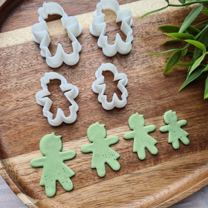 May include: A set of five white plastic cookie cutters shaped like people, along with five green fondant cookies cut out with the same shapes. The cutters and cookies are arranged on a wooden surface.