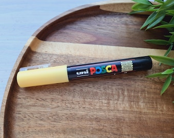 May include: A yellow Posca paint marker with black lettering that reads "uni POSCA" and lists the colors available in the marker set. The marker is on a wooden surface.