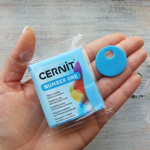 Könnte beinhalten: Eine blaue Packung CERNIT Number One Modelliermasse mit Porzellanglanz. Die Packung befindet sich in der Hand einer Person, und daneben liegt ein kleiner blauer Kreis aus Ton.