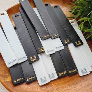 May include: An assortment of black and white plastic tools, each marked with measurements in millimetres, displayed on a wooden tray. The tools are rectangular with a hole at the top. Gold lettering indicates sizes from 2mm to 6mm.