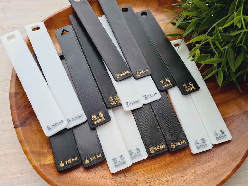 Peut inclure: Ensemble d'outils en plastique noir et blanc avec des mesures en millim&egrave;tres, dispos&eacute;s sur un plateau en bois. Les outils sont rectangulaires avec un trou en haut. Des lettres dor&eacute;es indiquent les tailles de 2 mm &agrave; 6 mm.