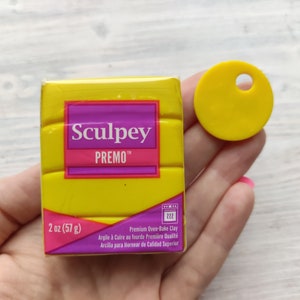 Puede incluir: Un paquete amarillo de 57 gramos de arcilla para hornear Sculpey Premo. El paquete tiene un diseño amarillo y morado con el nombre de la marca y el nombre del producto impresos en él. También se muestra un pequeño círculo amarillo con un agujero en el centro.