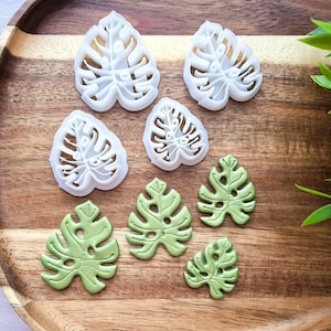 Puede incluir: Un juego de siete cortadores de galletas con forma de hojas de monstera. Los cortadores están hechos de plástico blanco y están dispuestos en una bandeja de madera. Hay cuatro cortadores más grandes y tres más pequeños. Los cortadores son perfectos para hacer galletas, fondant u otras decoraciones.