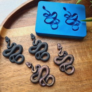 Puede incluir: Cuatro pendientes de resina de serpiente negra e iridiscente. Los pendientes tienen forma de serpiente enroscada. Los pendientes se muestran sobre una superficie de madera junto a un molde de silicona azul.