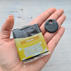 May include: A package of Cernit Pearl polymer clay with a small, round, gray pendant with a hole. The package is yellow and black with text in multiple languages. The pendant is approximately 2.5 cm in diameter.