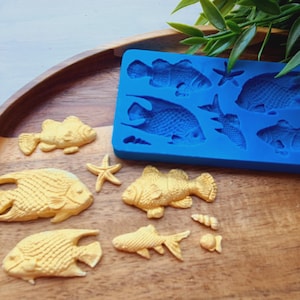 May include: A rectangular blue silicone mold featuring recessed shapes of various fish, a starfish, and a seashell. Beside the mold are several gold-colored, three-dimensional fish, a starfish, and small shell figures cast from the mold.