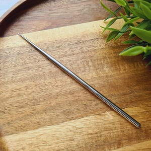 Puede incluir: Un palillo de pelo de metal plateado, con un extremo puntiagudo, descansa sobre una bandeja de madera. La bandeja tiene forma redonda y un acabado de vetas de madera natural. Ramitas de hojas verdes en el fondo.