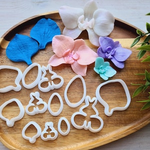 May include: A set of 14 white plastic cookie cutters shaped like orchids and leaves on a wooden surface. The cutters are arranged in a decorative pattern.
