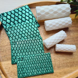 Op de afbeelding: Een houten dienblad toont turquoise vierkanten met schubbenpatroon en witte cilinders met honingraattextuur. De turquoise stukken zijn opgestapeld, de witte cilinders zijn verspreid. Een takje groen loof is zichtbaar.