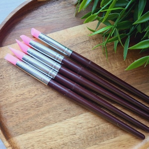Könnte beinhalten: Ein Set aus fünf Kunstwerkzeugen mit rosa Silikonspitzen, silbernen Metallzwingen und dunkelbraunen Holzgriffen. Die Werkzeuge sind auf einem Holztablett angeordnet, mit grünem Laub im Hintergrund.