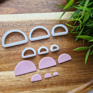 Puede incluir: Un conjunto de 10 cortadores de galletas de plástico blanco de diferentes tamaños, con forma de semicírculos y arcos. Los cortadores están dispuestos sobre una superficie de madera.