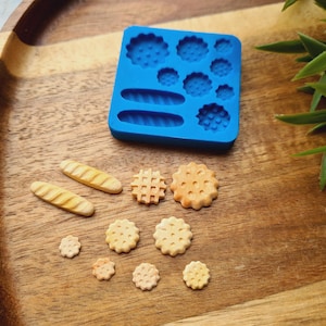 Può includere: Stampo in silicone blu con varie forme per realizzare cibo in miniatura, tra cui pane, biscotti e pasticcini. Lo stampo è mostrato con diversi articoli di cibo in miniatura finiti, tra cui un waffle, un biscotto e due grissini.