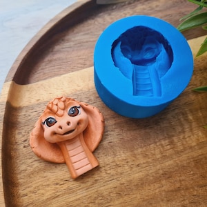 Könnte beinhalten: Eine Silikonform zum Herstellen eines kleinen, orangefarbenen Drachenkopfes mit großen Augen. Die Form ist blau und hat eine detaillierte Prägung des Drachenkopfes.