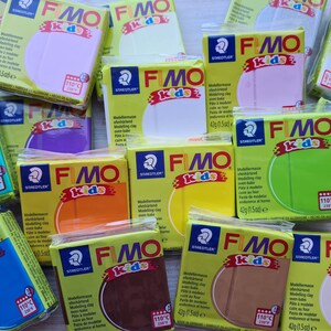 May include: An assortment of 12 colourful modelling clay bars in various colours, including yellow, orange, brown, green, blue, purple, and white. Each bar is labelled "FIMO kids" and has a Staedtler logo. The clay is oven-bake and suitable for crafting.