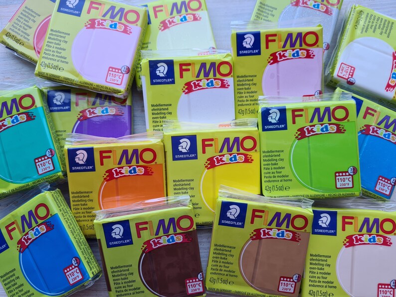 May include: An assortment of 12 colourful modelling clay bars in various colours, including yellow, orange, brown, green, blue, purple, and white. Each bar is labelled "FIMO kids" and has a Staedtler logo. The clay is oven-bake and suitable for crafting.