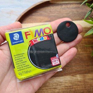 May include: A pack of black FIMO Kids modelling clay with the text "STAEDTLER FIMO kids" and "Modelliermasse ofenh&auml;rtend Modelling clay oven-bake P&acirc;te &agrave; modeler cuire au four Pasta de modelar endurence al horno 42g (1.5 oz) e MADE IN GERMANY FABRIQUE EN ALLEMAGNE 110&deg;C 230&deg;F". A black circle with a hole in the centre is in the background.