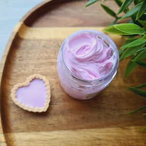 Può includere: Un barattolo di vetro trasparente riempito con un vortice di burro corporeo montato rosa chiaro, appoggiato su un vassoio di legno con un biscotto a forma di cuore.