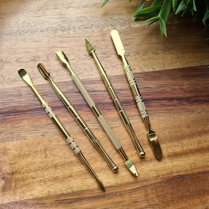 May include: A set of five gold-colored metal tools with textured grips, each featuring distinct tips including rounded, pointed, and flat spatula ends. These slender implements are arranged diagonally, showcasing a polished finish and varied shapes.
