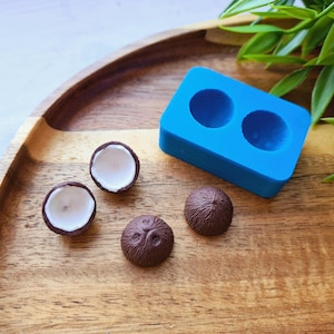Peut inclure: Un moule en silicone bleu avec deux cavités en forme de noix de coco. Il y a quatre moitiés de noix de coco brunes et deux noix de coco entières sur une surface en bois.