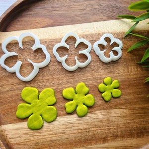 Puede incluir: Cuatro cortadores de galletas de plástico blanco en forma de flor de diferentes tamaños, y tres flores de fondant verde, sobre una superficie de madera.
