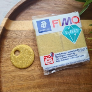 May include: A package of FIMO Effect modeling clay, 57g (2 oz), in a gold color, next to a finished, round, gold-colored clay pendant with a hole. The package has text in multiple languages, including "oven-bake".
