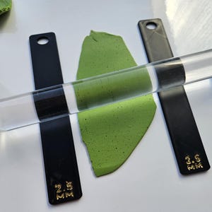 Peut inclure: Un rouleau en acrylique transparent est pos&eacute; sur une feuille d'argile verte, flanqu&eacute; de deux guides en acrylique noir. Les guides sont marqu&eacute;s d'une inscription dor&eacute;e indiquant une &eacute;paisseur de 2,5 mm et 3,5 mm. Outils de cr&eacute;ation.
