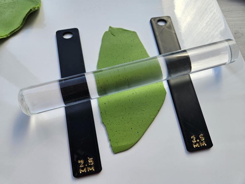 Peut inclure: Un rouleau en acrylique transparent est pos&eacute; sur une feuille d'argile verte, flanqu&eacute; de deux guides en acrylique noir. Les guides sont marqu&eacute;s d'une inscription dor&eacute;e indiquant une &eacute;paisseur de 2,5 mm et 3,5 mm. Outils de cr&eacute;ation.