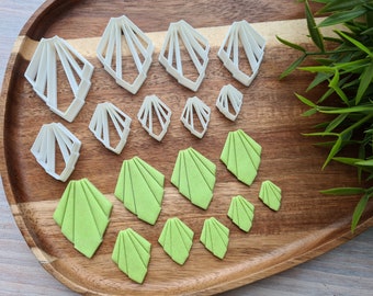 May include: A set of 12 white plastic cookie cutters in various sizes and shapes, featuring a geometric fan design. The cutters are arranged on a wooden surface with green fondant cutouts.