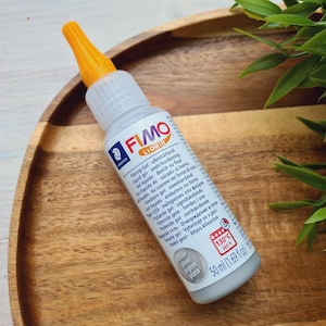 May include: A silver FIMO Liquid bottle with a white label and orange cap. The label has the text "FIMO LIQUID" and instructions for using the product. The bottle is 50 ml (1.69 fl oz).