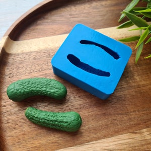 Peut inclure: Un moule en silicone bleu vif présentant deux cavités incurvées et allongées conçues pour le moulage. Deux petits objets texturés en forme de cornichon vert foncé sont placés à côté du moule, illustrant le résultat final du moulage.