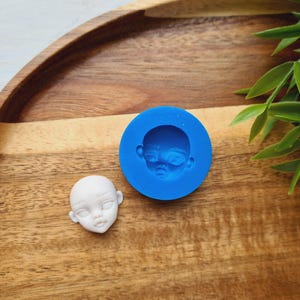 Puede incluir: Un molde de silicona azul para hacer una cara de muñeca, con una cara de muñeca blanca hecha del molde al lado.